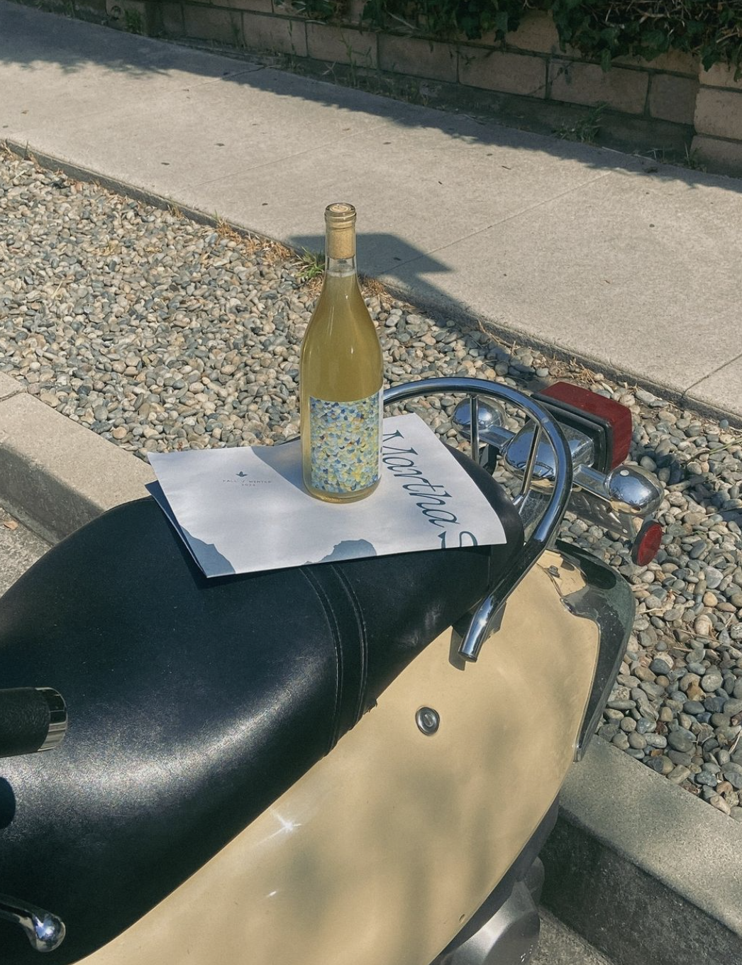 a newspaper and a bottle of wine sit upon the seat of a vespa scooter at golden hour